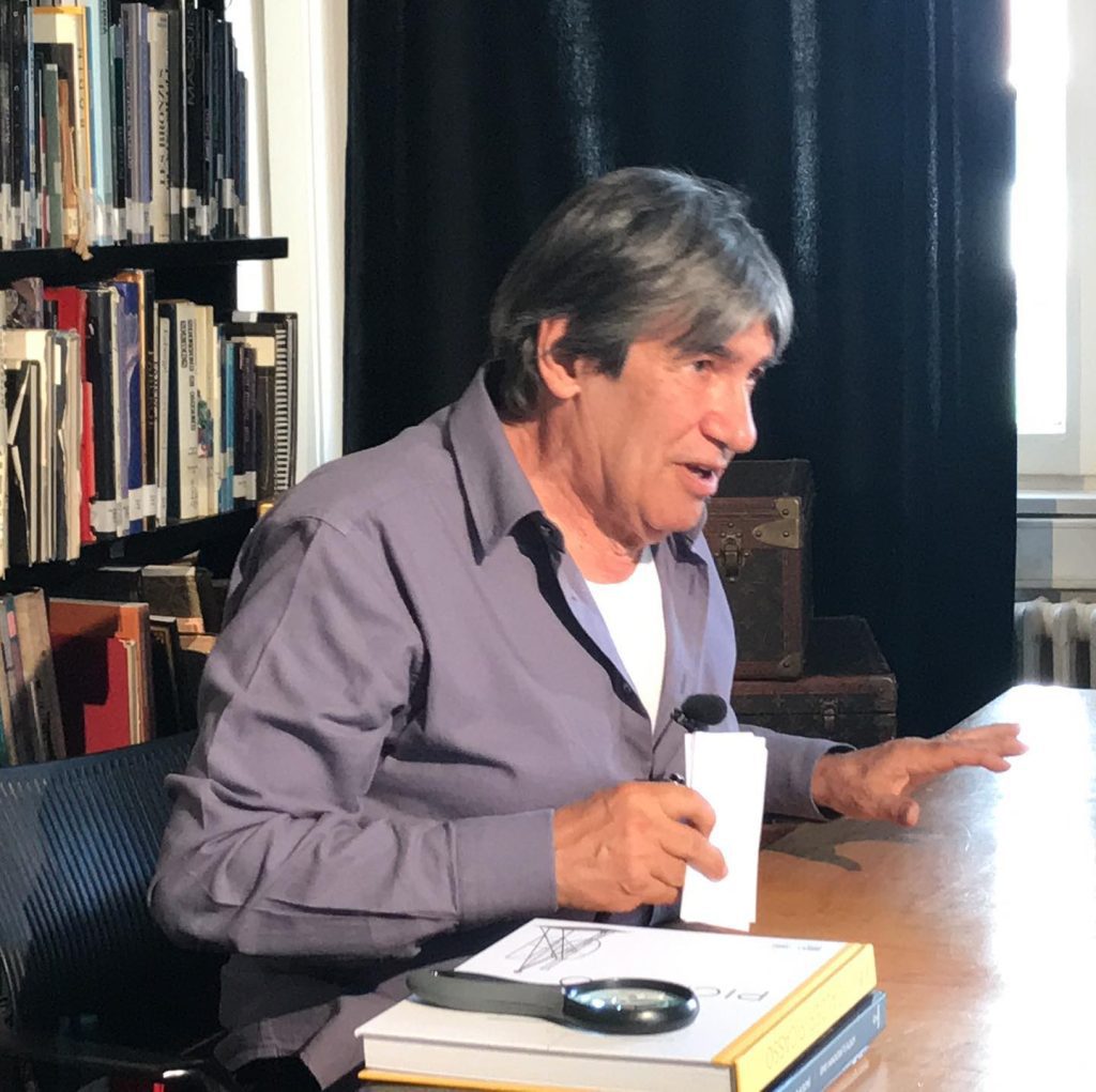A man sitting at a table, with a book laid out next to him, appearing engaged in a conversation.